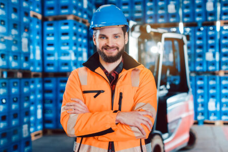 Mitarbeiter in orangefarbener Warnjacke und blauem Schutzhelm steht im Getränkelager vor gestapelten Kisten; im Hintergrund ein Gabelstapler.
