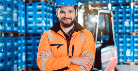 Mitarbeiter in orangefarbener Warnjacke und blauem Schutzhelm steht im Getränkelager vor gestapelten Kisten; im Hintergrund ein Gabelstapler.
