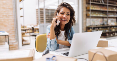 Mitarbeiterin im Büro telefoniert und berät Kunden; am Arbeitsplatz stehen Laptop und Pakete – Service und Bestellannahme.